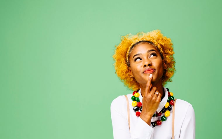 A woman standing in front of a green while while thinking about a decision with her finger resting on her chin.