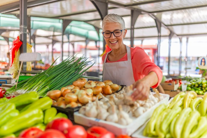 A woman standing behind her food stall at a farmers market and rearranging vegetables.