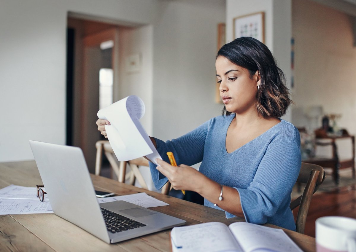 Woman looking over her bills with laptop and notepad at kitchen table.