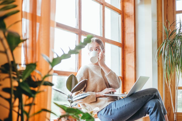 A woman sitting in a suy window seat at home while drinking coffee and typing on her laptop.