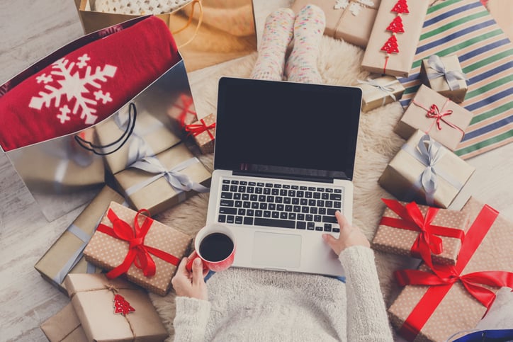 A woman sitting on the floor holding a coffee with her laptop open and surrounded by wrapped gifts.