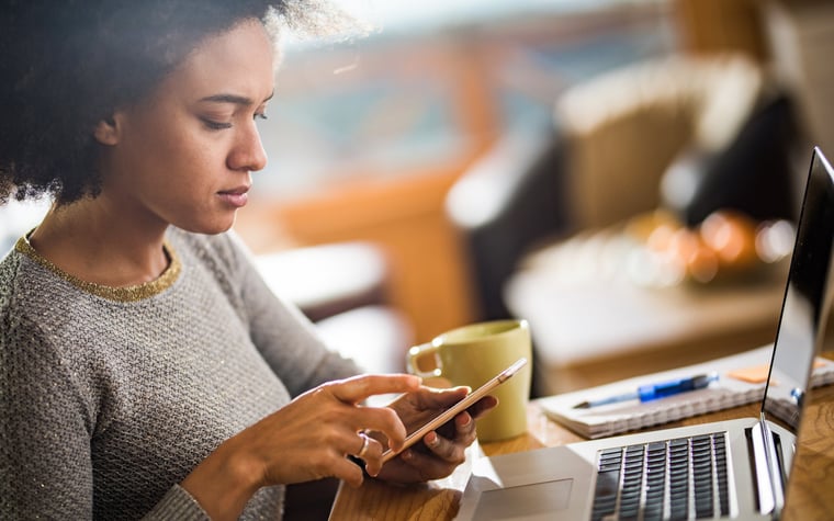 A woman dialing a number on her phone and sitting at a table with her laptop.