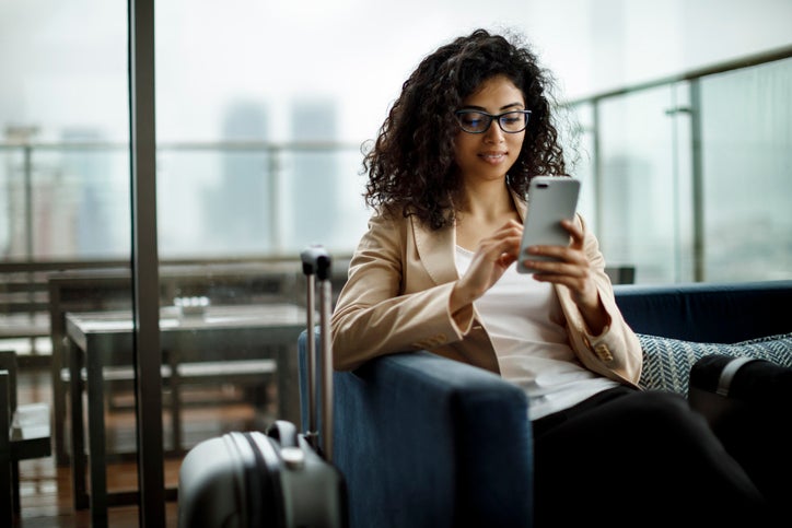 Woman sitting next to suitcase in airport lounge and looking at mobile phone.