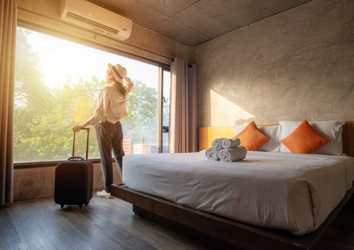 A woman standing next to her suitcase and looking out the window of her sunny hotel room.
