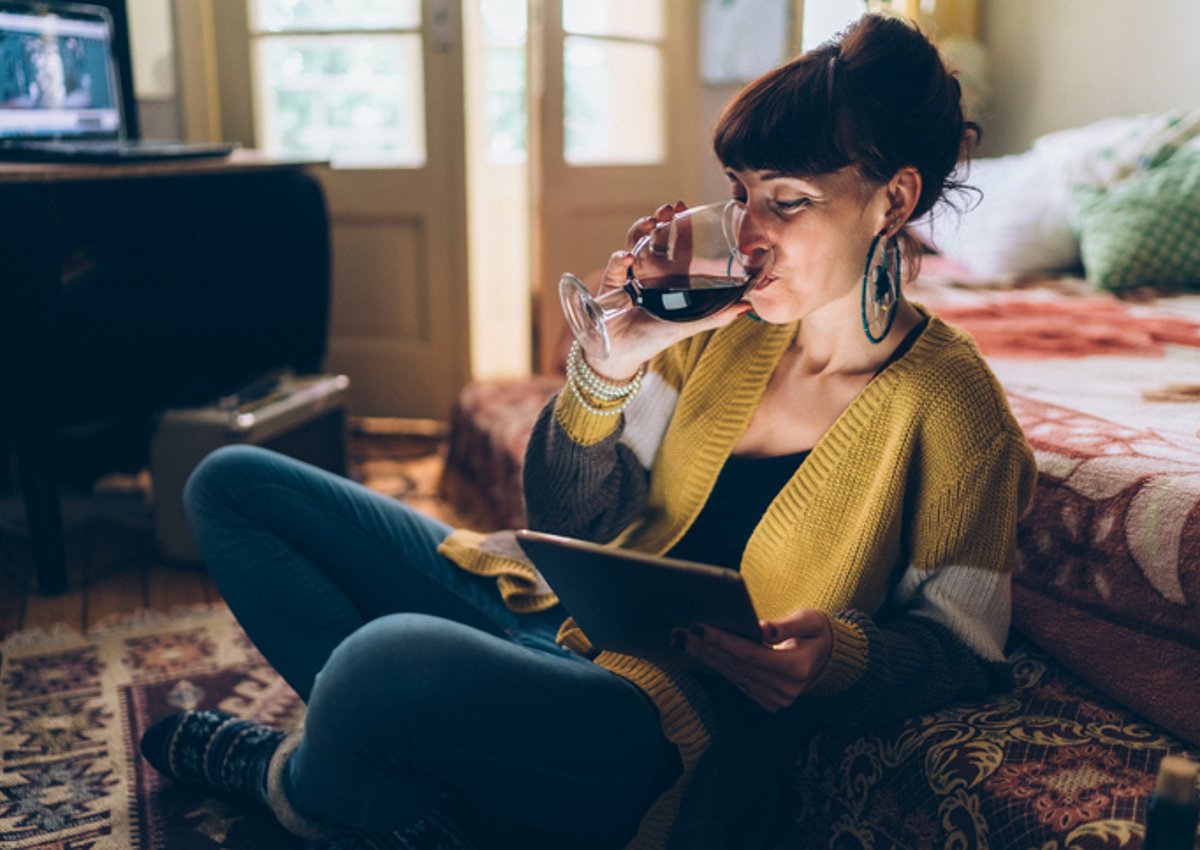 A woman sitting on the floor drinking wine while watching a movie on her tablet.