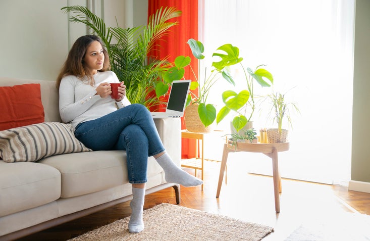 Woman sitting on sofa with hot drink and laptop.