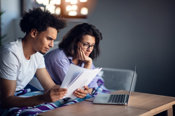A worried-looking man and woman sitting on their couch and going through paper bills with a laptop open on the coffee table.