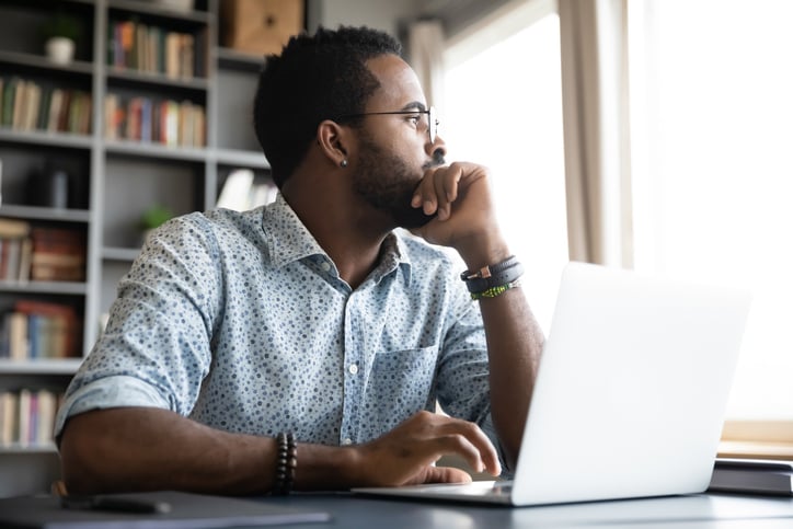 A person sitting at a desk in front of an open laptop and glancing out the window to his left.