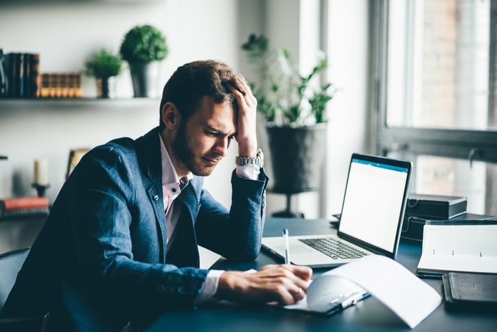 A man sitting at his office desk and looking stressed with his hand on his forehead while he fills in paperwork.