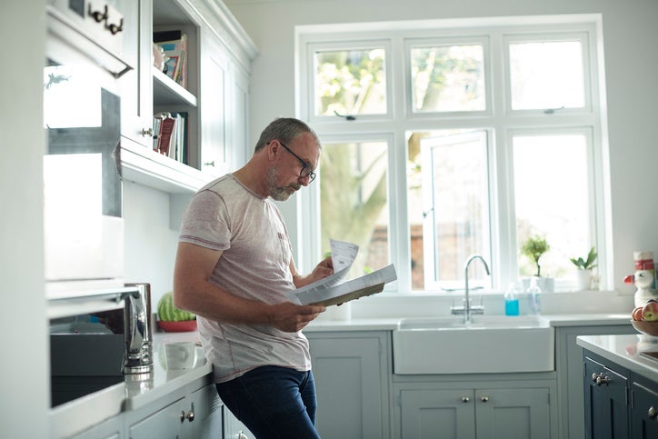 A worried-looking man standing in his kitchen and reading through paper bills in his hands.
