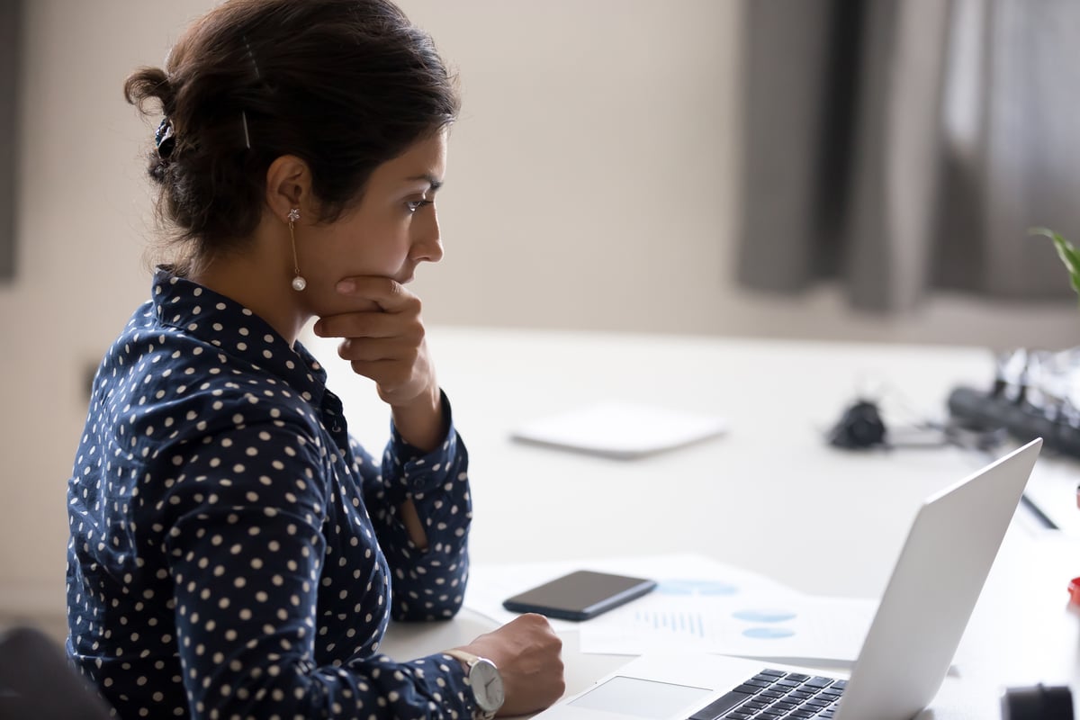 Une femme inquiète avec sa main sur son menton assise à un bureau avec des papiers et son ordinateur portable.