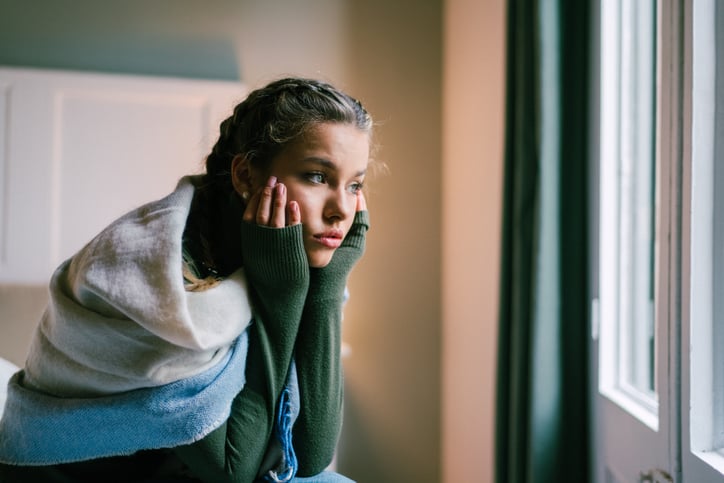 A worried young woman sitting on the edge of her bed with her chin in her hands and a blanket around her shoulders.