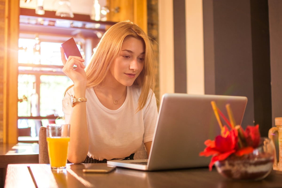 A young woman sitting at a sunny dining room table in front of her laptop and holding a credit card.