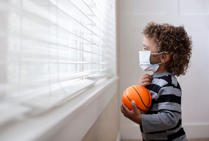 A young boy holding a small basketball and wearing a medical mask while looking forlornly out the window.