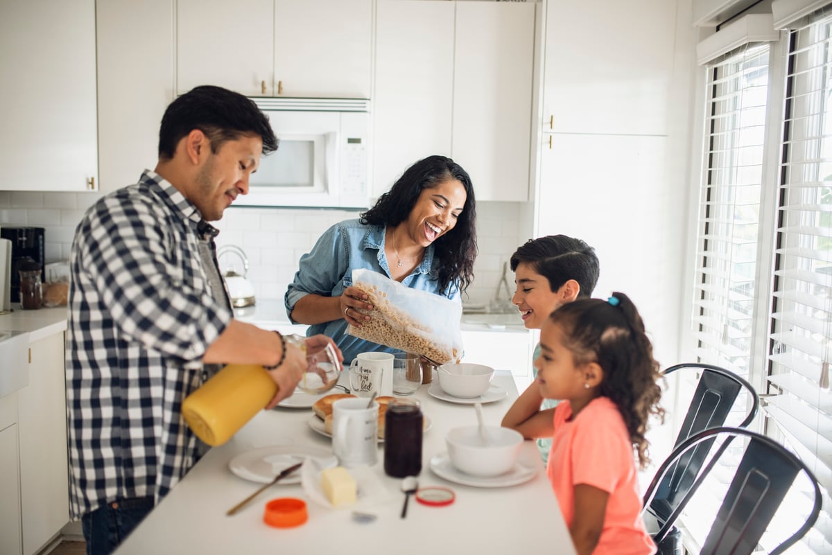 Two parents making breakfast for their young kids sitting in the kitchen.