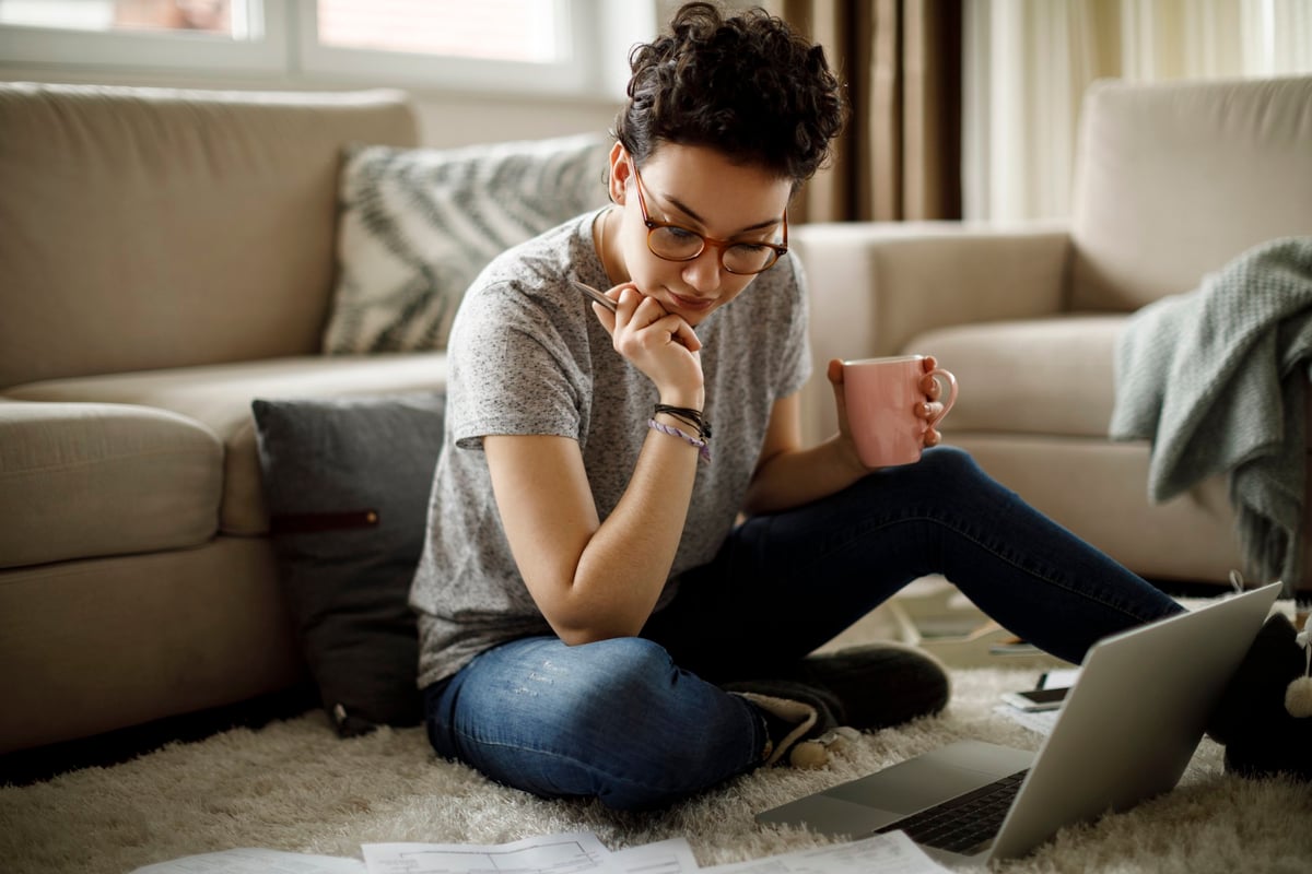 A young woman sitting on the floor of her living room with her laptop and bills around her.