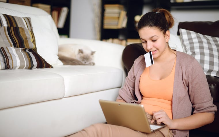 A young woman shops on her laptop holding her credit card while her cat lounges in the background