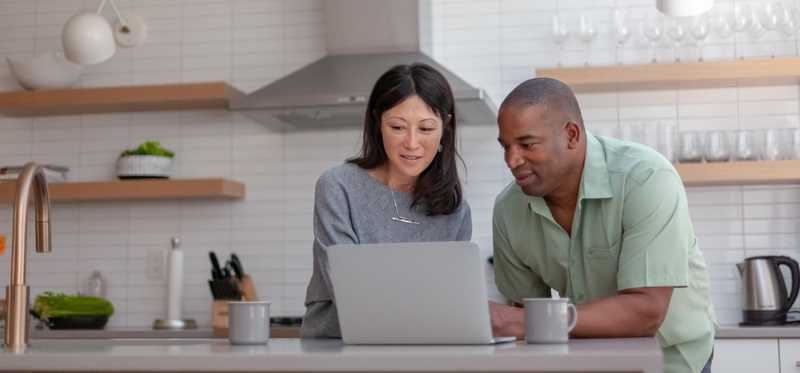 Two people talk while looking at laptop in kitchen.