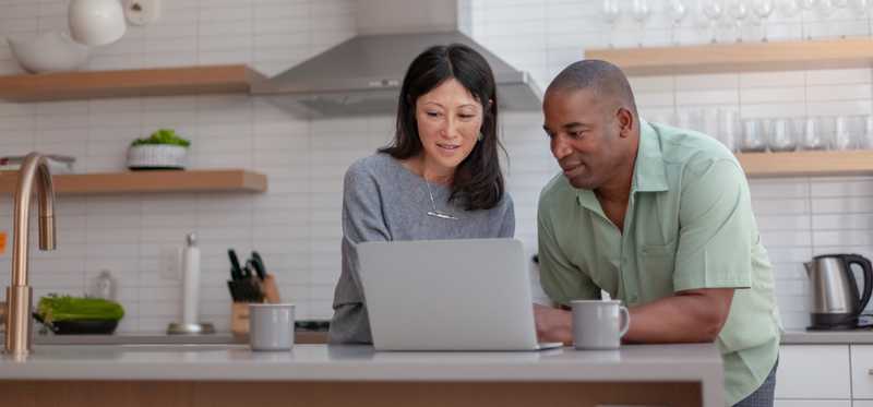 Two people talk while looking at laptop in kitchen.