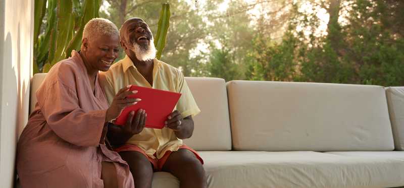 Two people smiling and laughing while looking at tablet and sitting on couch outdoors.