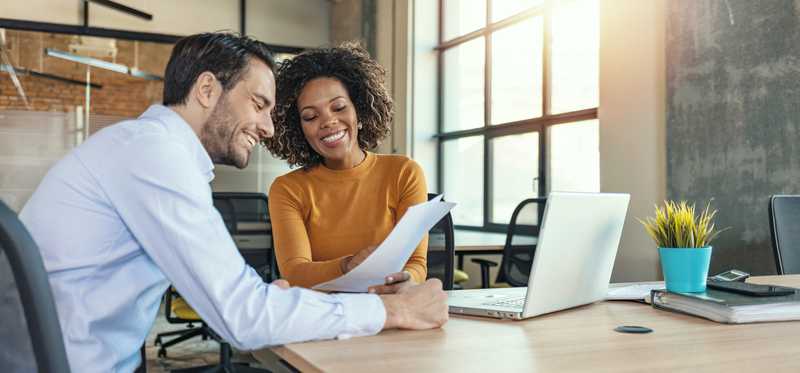 Two people seated in office while looking at paperwork and smiling.