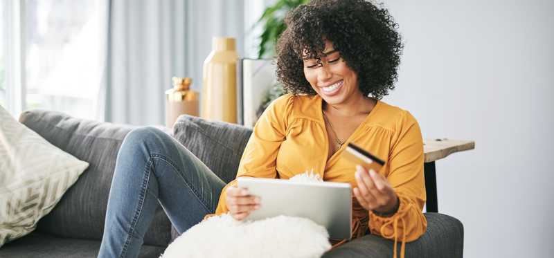 Person sitting on couch at home holds credit card and laptop.