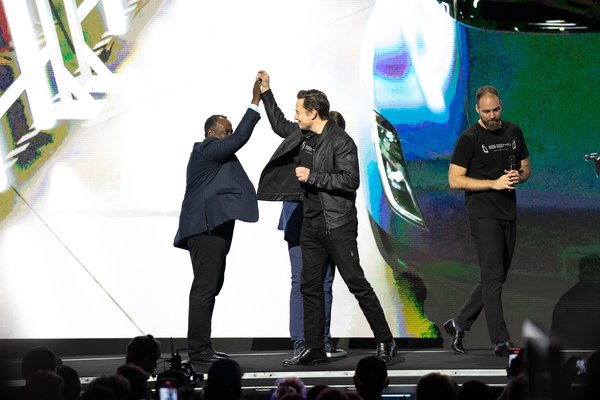Elon Musk (middle) stands on stage and joins hands with another event participant.