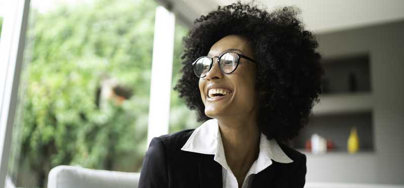 Adult wearing professional attire smiles while sitting on couch in front of window.