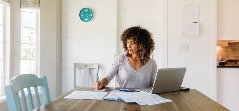 Person takes notes at dining table, surrounded by papers and laptop.