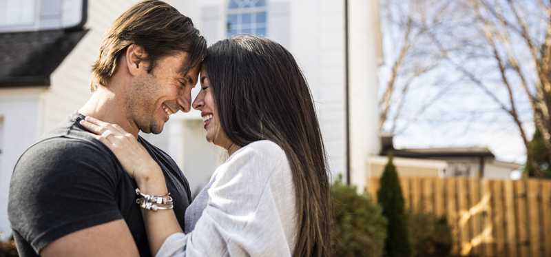 Adult couple embraces in front yard of home.