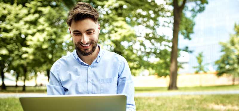 Smiling adult sits outside in the park with laptop.