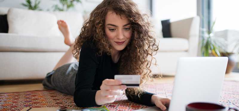 Person lying on floor is looking at credit card while using laptop.