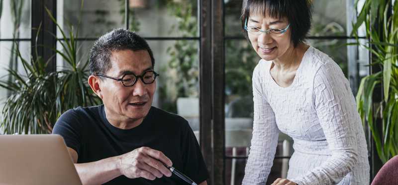 Two people reviewing paperwork on desk by glass doors and plants.