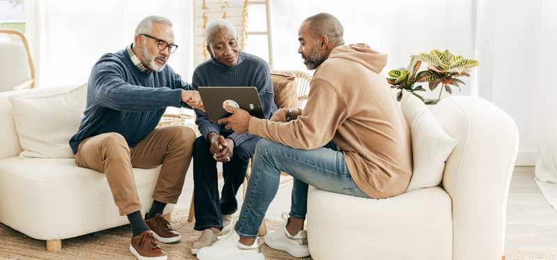Two people explain information on laptop to another person while sitting on the couch at home.