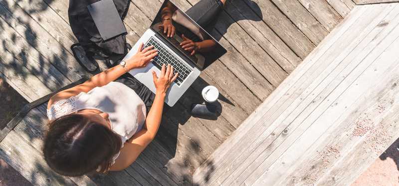 Person sitting in the shade on a wooden deck and typing on a laptop.