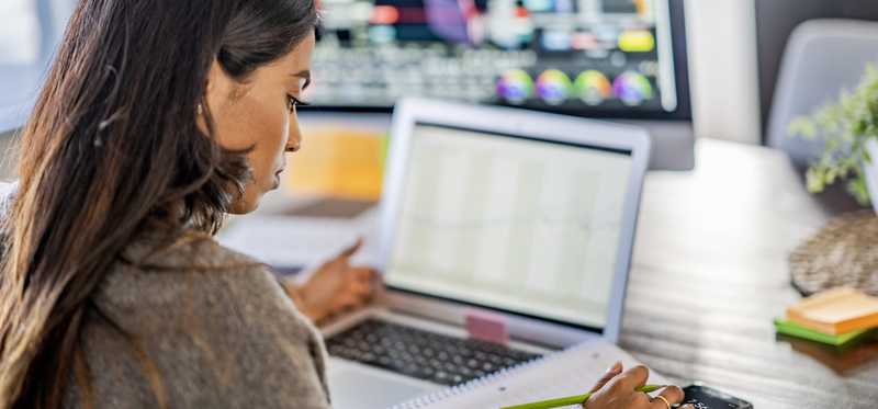 Adult uses calculator in front of laptop next to a larger monitor with charts.