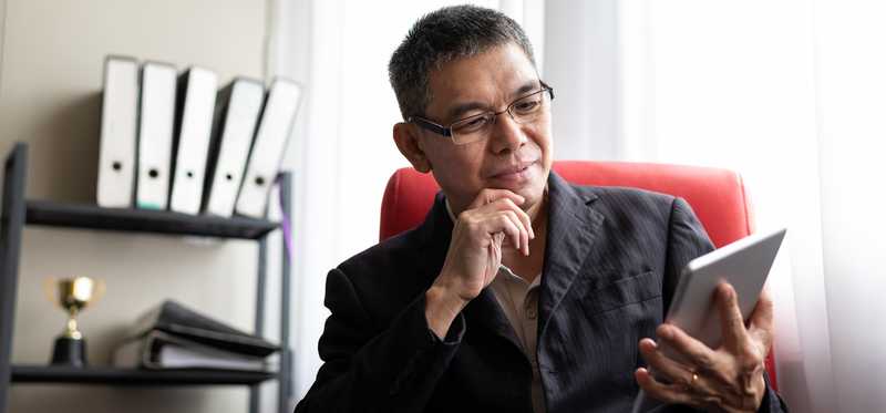 Person wearing professional attire and glasses looks thoughtfully at tablet.