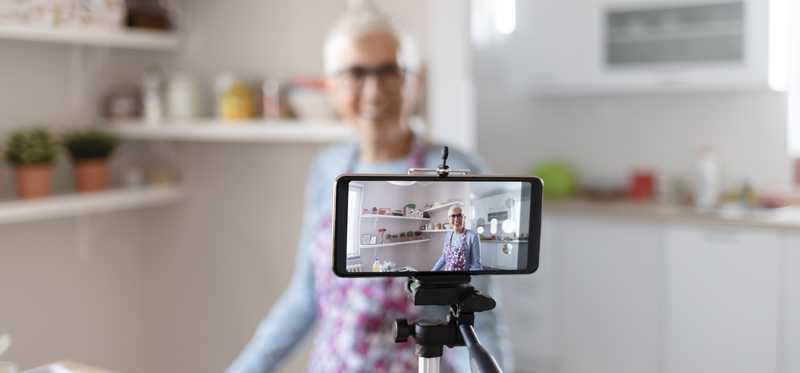 Smiling older woman in kitchen as seen on phone screen held on tripod