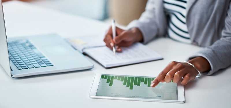 Woman refers to chart displayed on tablet while writing in notebook.