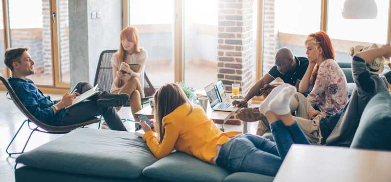 Group of people sit in living room together with laptops and smartphones.