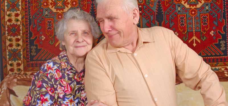 Elderly woman holds on to the arm of her husband as they sit on the couch together.