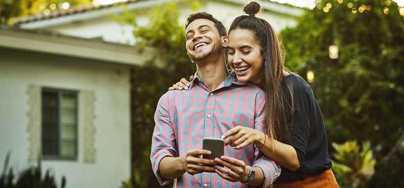 Two people laughing while looking at phone outside house.
