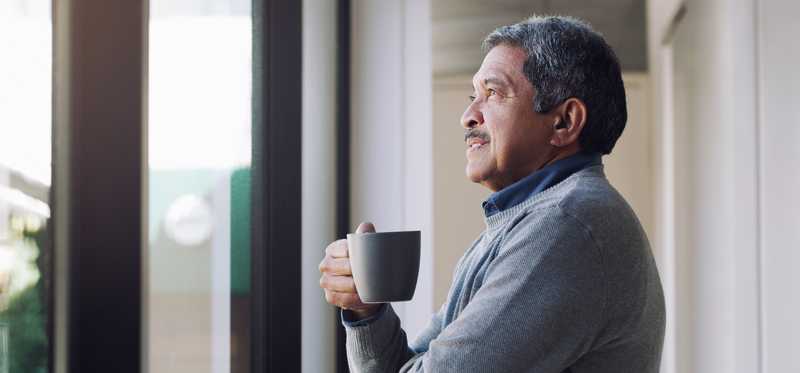 Person looks out window thoughtfully while holding coffee mug.
