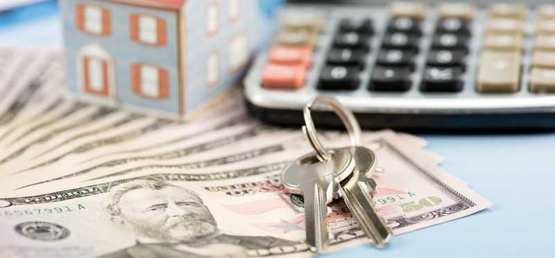 A small model house sits atop cash next to house keys and a calculator.
