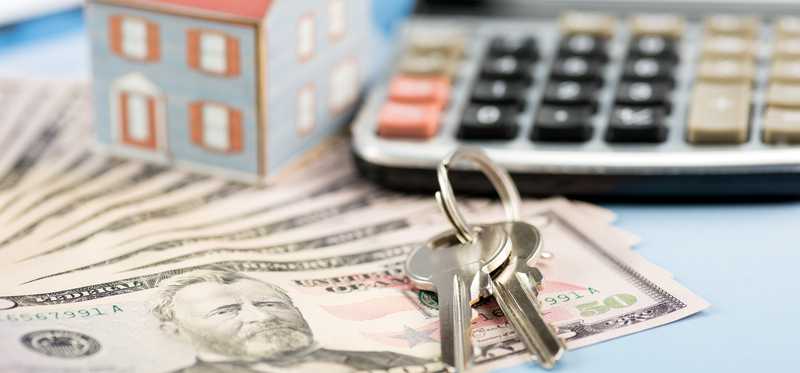 A small model house sits atop cash next to house keys and a calculator.