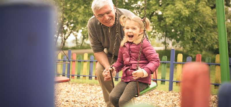 An elderly man pushes a toddler girl on a swing.