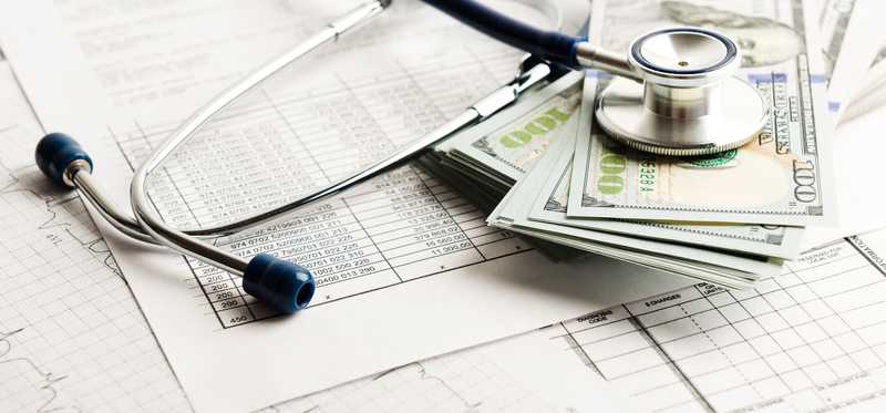 A stethoscope sits atop a pile of money and various paperwork showing data.