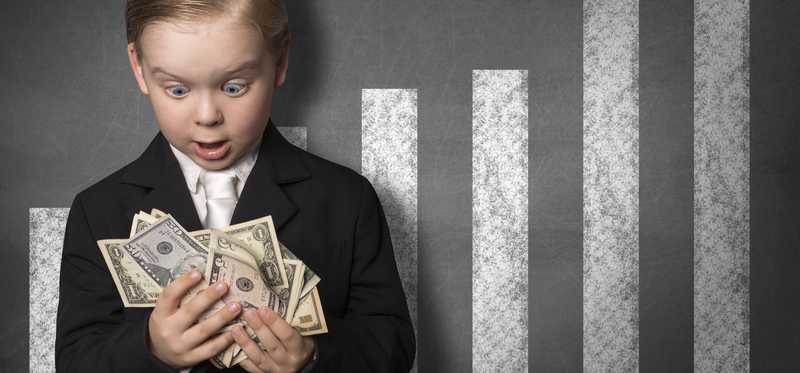A young boy is holding a lot of cash in his hands, looking amazed, standing in front of an upward-sloping bar graph on a blackboard.