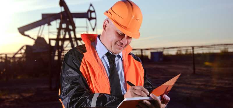 Man in orange hard hat and vest holding notebook in front of oil well.