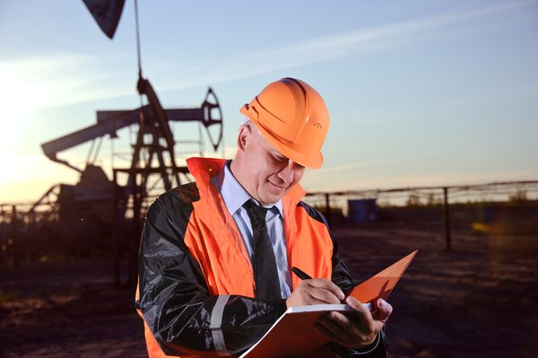 Man in orange hard hat and vest holding notebook in front of oil well.